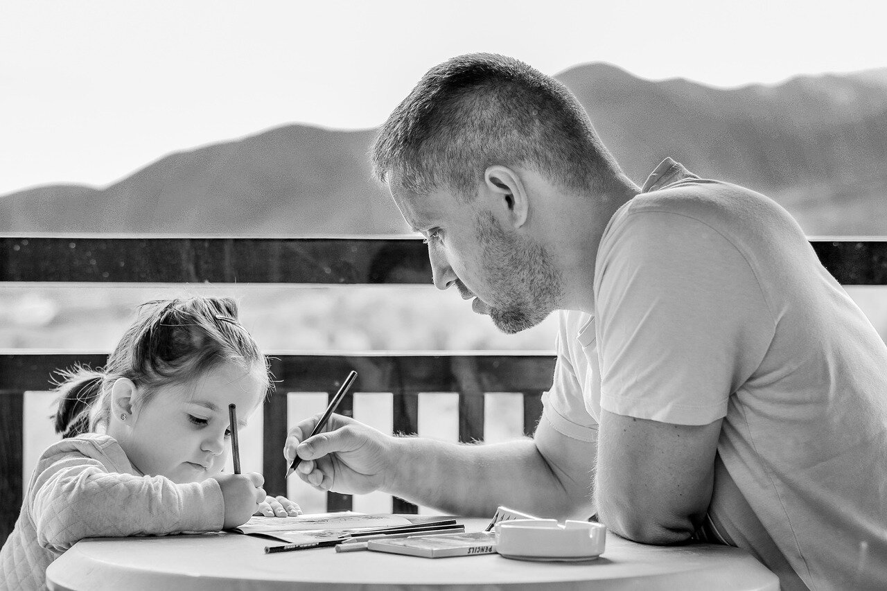 A parent works at a desk in a home office while a child plays on the floor nearby, illustrating the balance between professional duties and family life in a remote work setting.